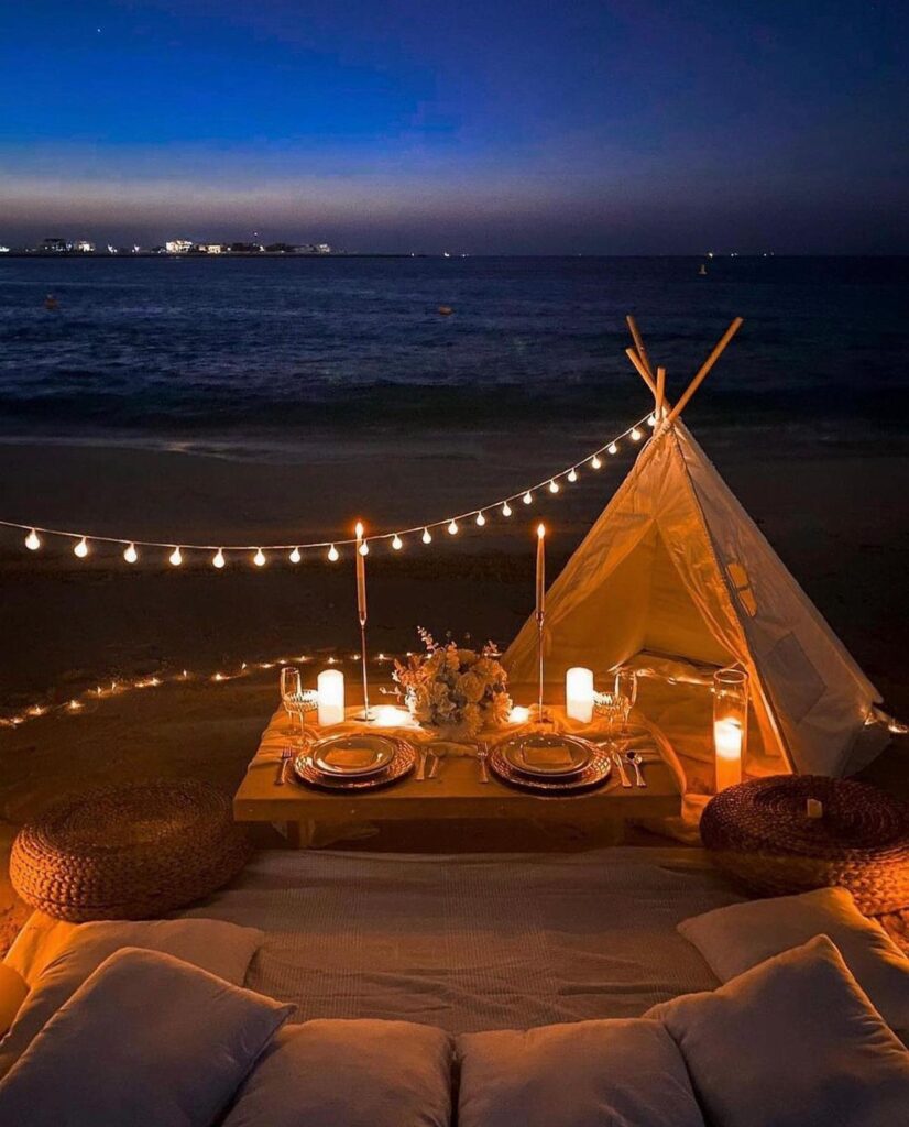 Romantic beach picnic setup with a teepee, string lights, and dinner for two.