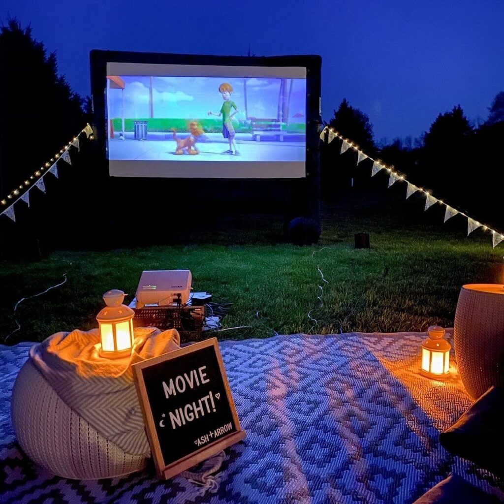 Outdoor movie screen with lanterns and blankets on a lawn at dusk.