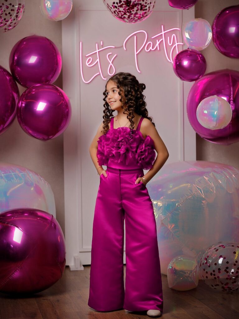 Girl in magenta satin ruffled top and wide-leg pants among pink balloons