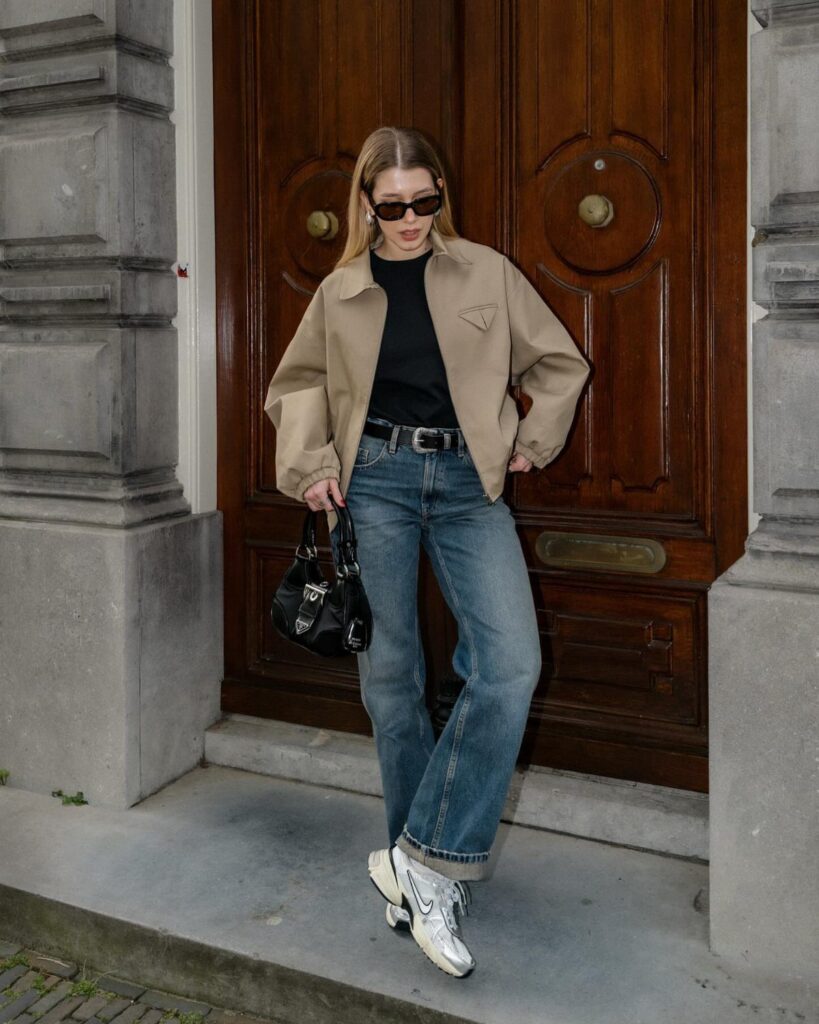 Woman in beige jacket and blue jeans posing in front of wooden doors.