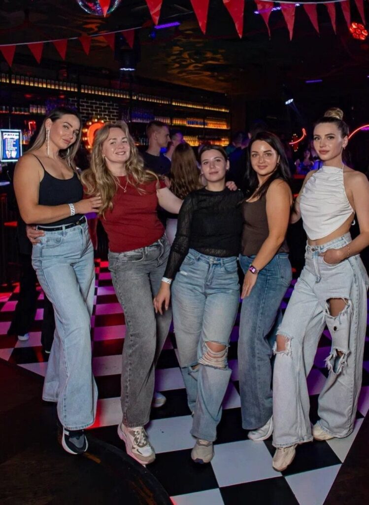 Five women in casual tops and denim jeans posing on a checkered dance floor.
