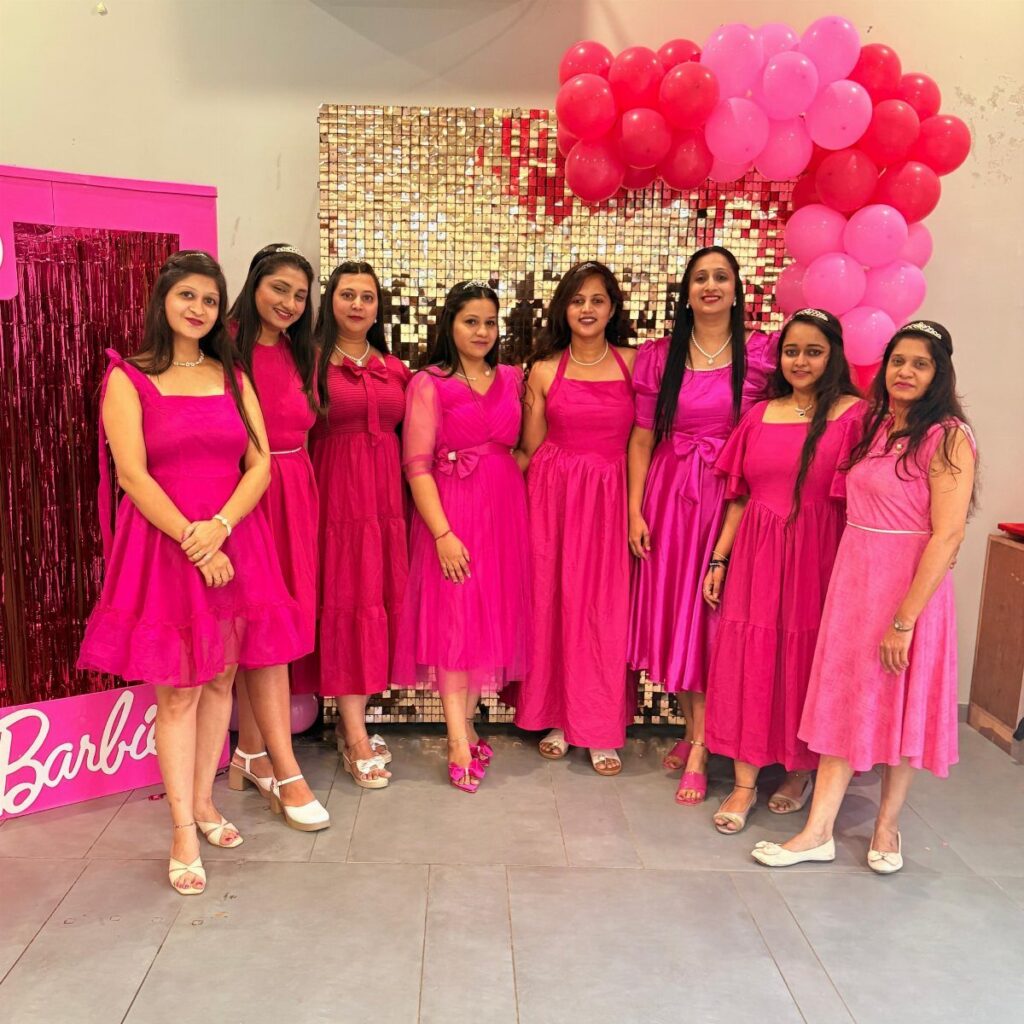 Eight women in pink dresses posing before a gold sequin wall and balloons.