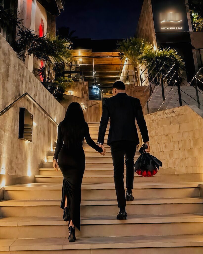 Couple in formal black attire walking up stairs, man holding red roses.