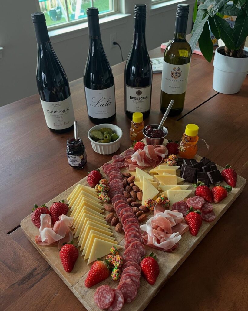 Wine bottles and a wooden cheese board with meats and fruit on a table.