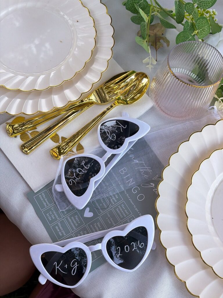 Heart-shaped sunglasses with initials and date beside gold cutlery and white plates.