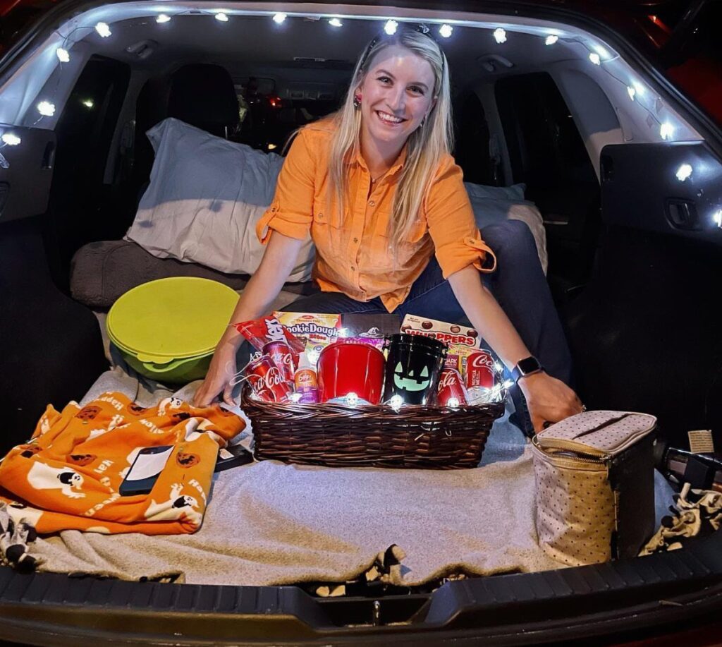 Woman sitting in a car trunk with blankets, snacks, and string lights.