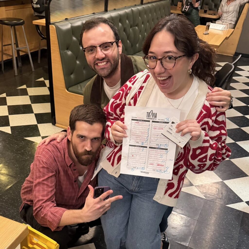Three friends posing in a diner, holding a trivia night answer sheet.