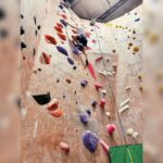Person climbing a tall indoor wall with colorful handholds and safety rope.