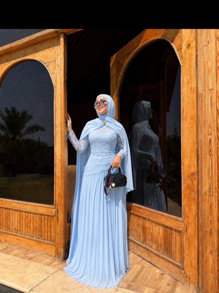 Woman in a light blue modest lace gown and hijab standing by wooden arches.