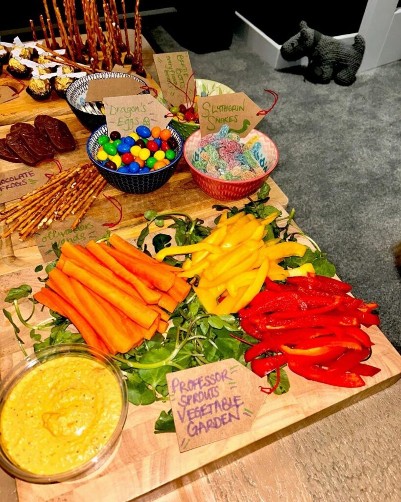 Assorted snacks including vegetable sticks, dip, and colorful candies on a wooden board.
