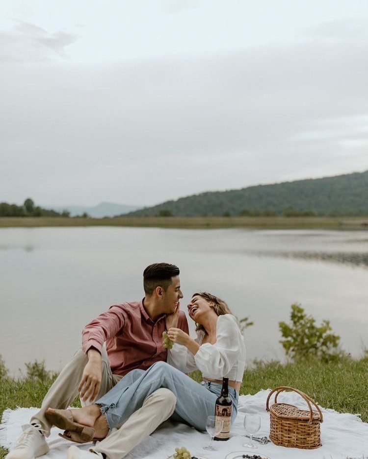 Couple laughing on a picnic blanket by a lake with wine and basket.