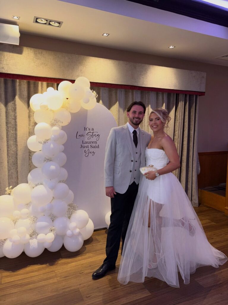 Couple posing in front of a white balloon arch and engagement announcement backdrop.