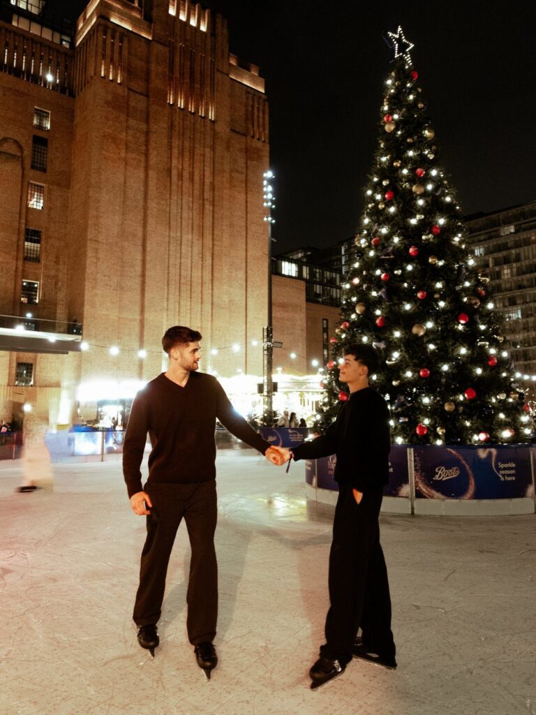 Two men holding hands while ice skating outdoors next to a large Christmas tree.